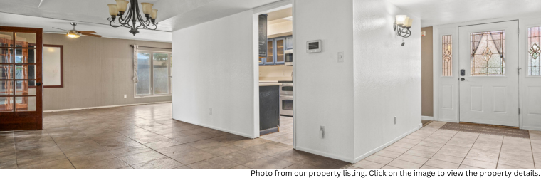 Interior of home with featuring living room and entry tan ceramic tiles and white and tan walls. Gives glimpse into kitchen.